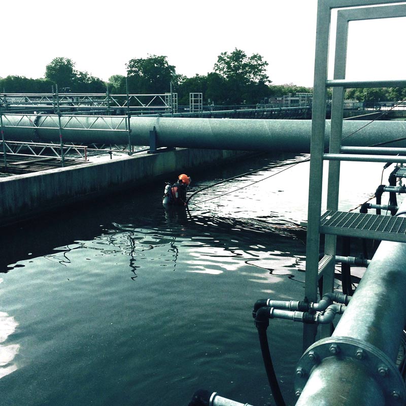 Ein Industrietaucher steht in voller Ausrüstung, knietief im Wasser begutachtet das Wasserbecken einer Industrieanlage